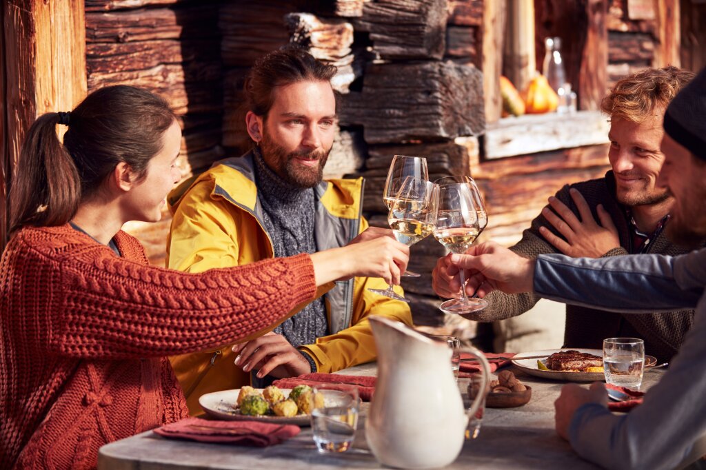 Foto: IDM Südtirol-Alto Adige/Anmi Freunde stoßen in einer Stube mit Wein an, auf dem Tisch Südtiroler Gerichte.